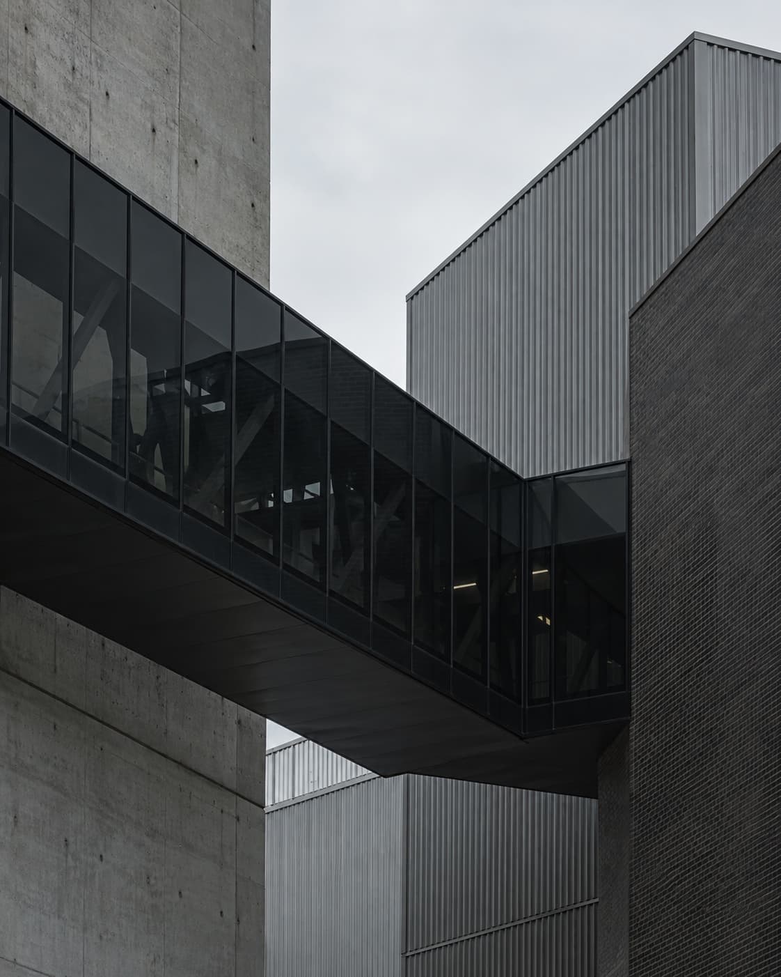 A dark enclosed bridge joining concrete, metal, and masonry buildings.